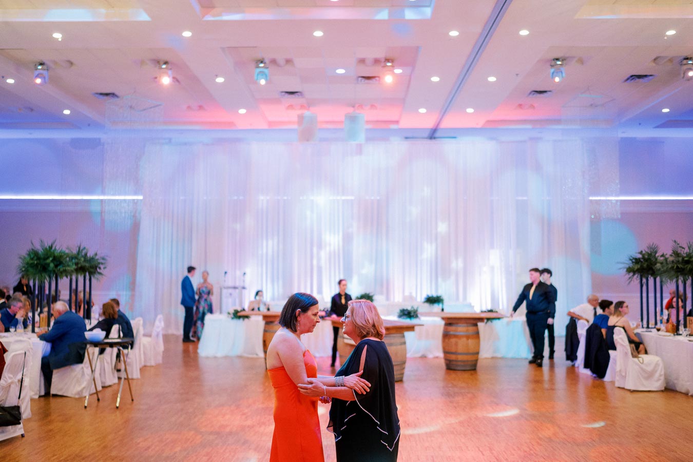 Two women converse on the dance floor at an elegant indoor event, surrounded by seated guests at decorated tables. The venue features sophisticated lighting, ceiling decor, and lush greenery, creating a warm and inviting atmosphere.