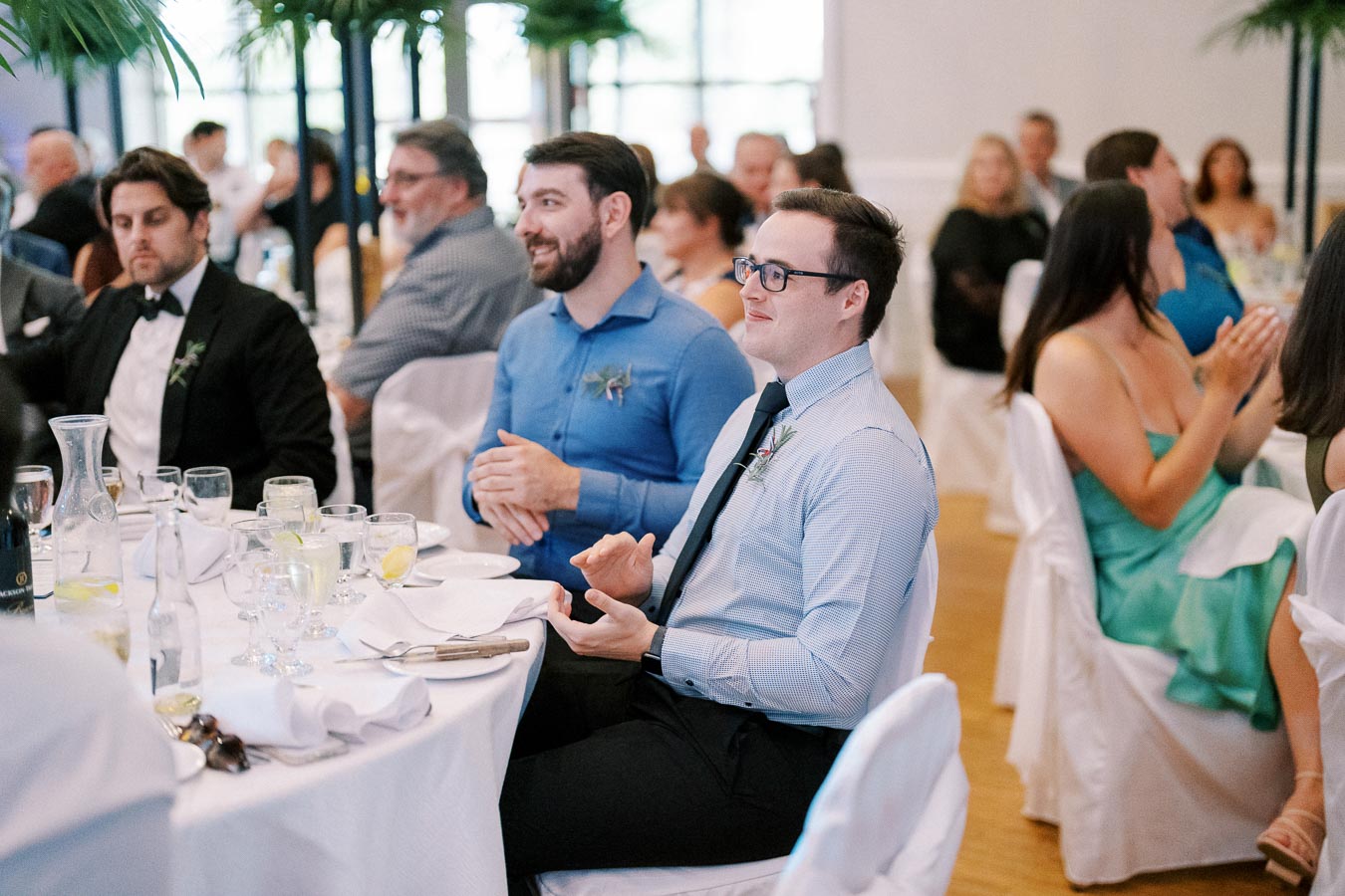 Guests sitting at a formal event table, dressed in elegant attire with glasses and plates set in front of them, participating and enjoying the gathering surrounded by other seated attendees.