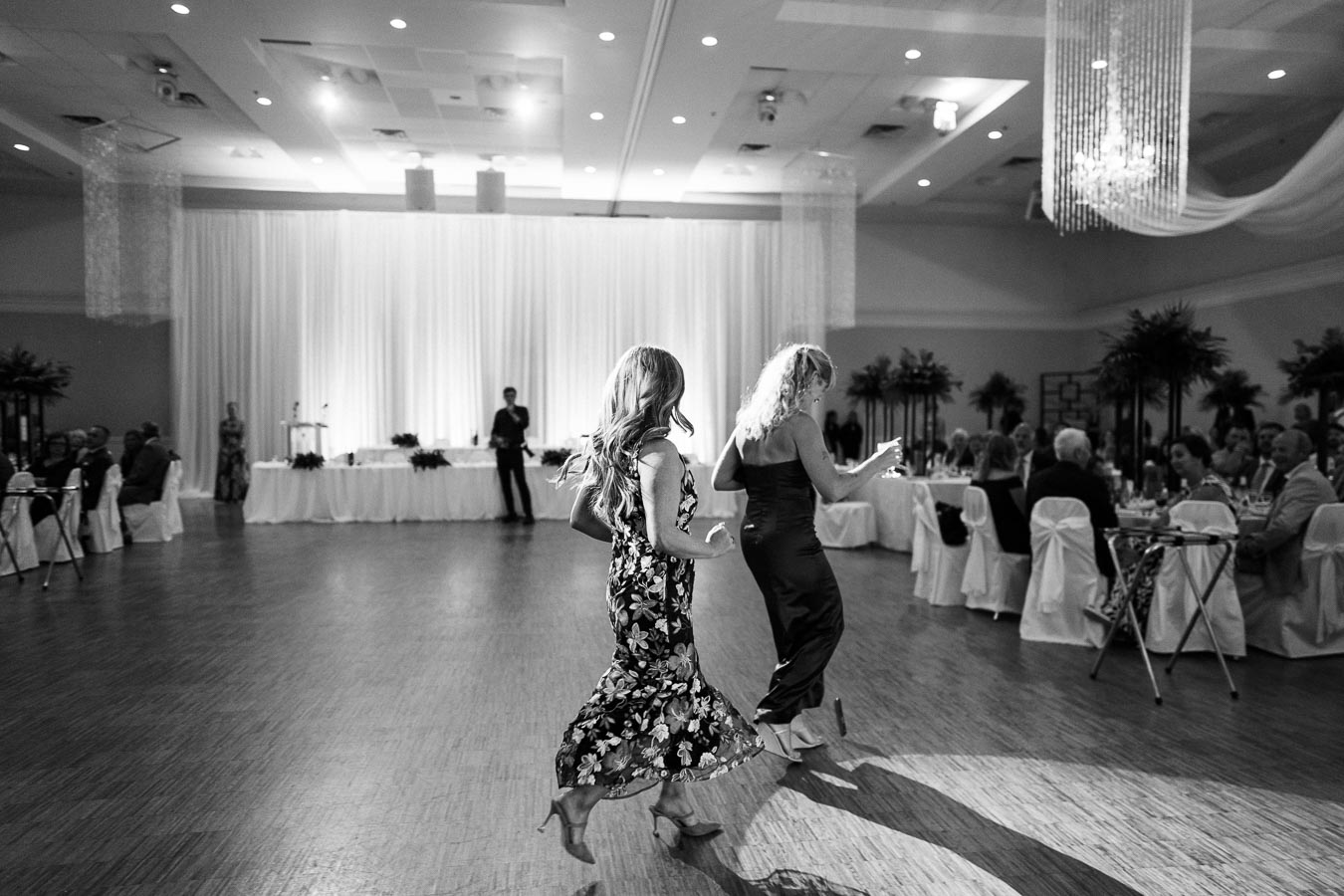 Two women dancing joyfully at a formal event in a banquet hall, with tables set for guests and elegant decor.