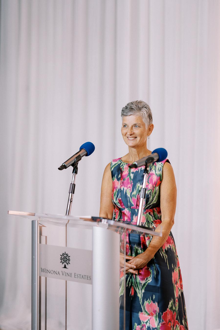 Elegant woman in a floral dress speaking at a podium with microphones at Winona Vine Estates event.