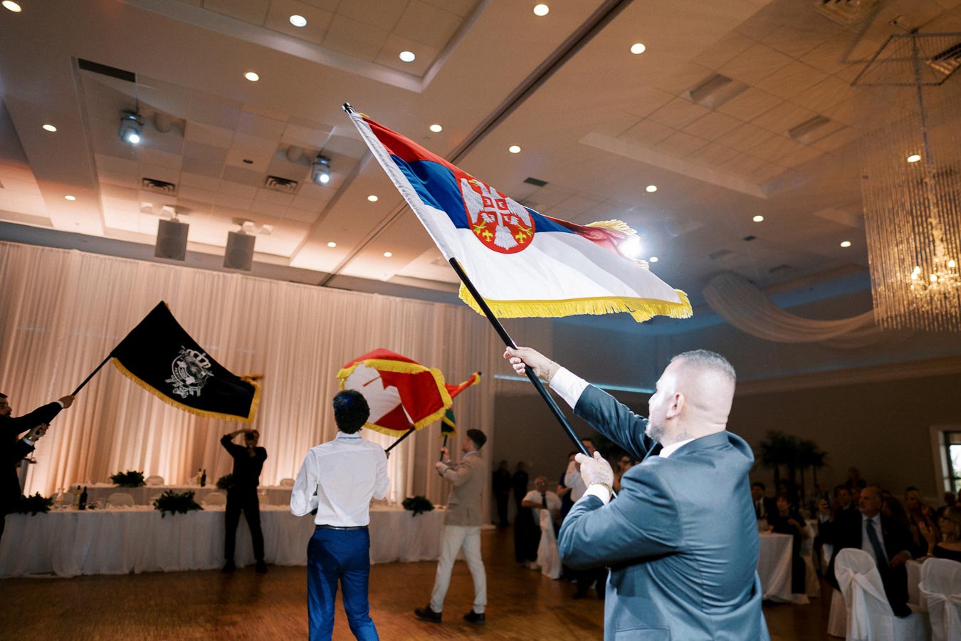 A person in formal attire proudly waving a Serbian flag indoors during a celebration, with other people holding different flags in the background. The event is set in a well-lit banquet hall with elegant decor.