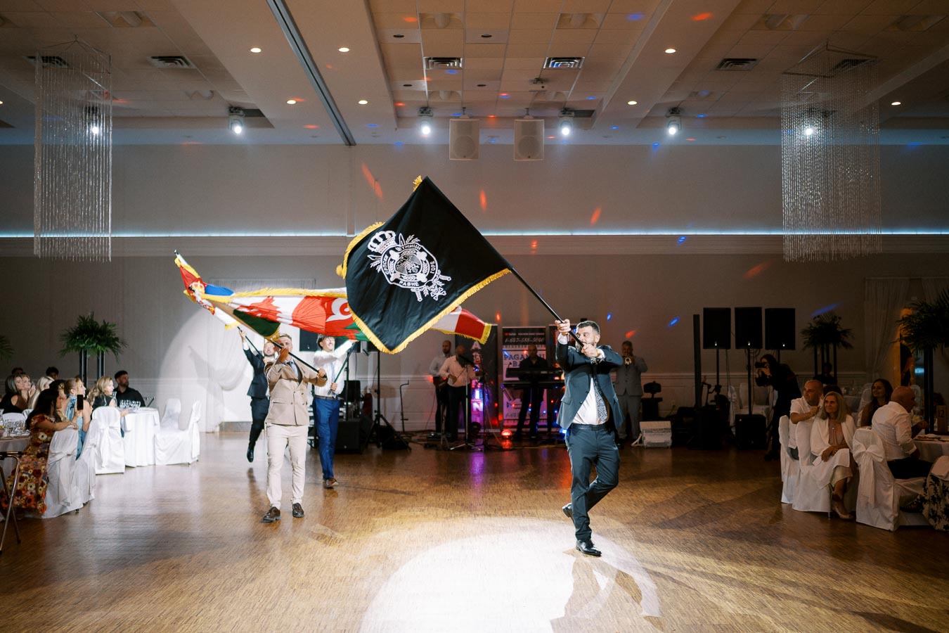 A dynamic scene in a well-lit banquet hall, featuring individuals marching with colorful flags, creating a lively and festive atmosphere during an indoor event.