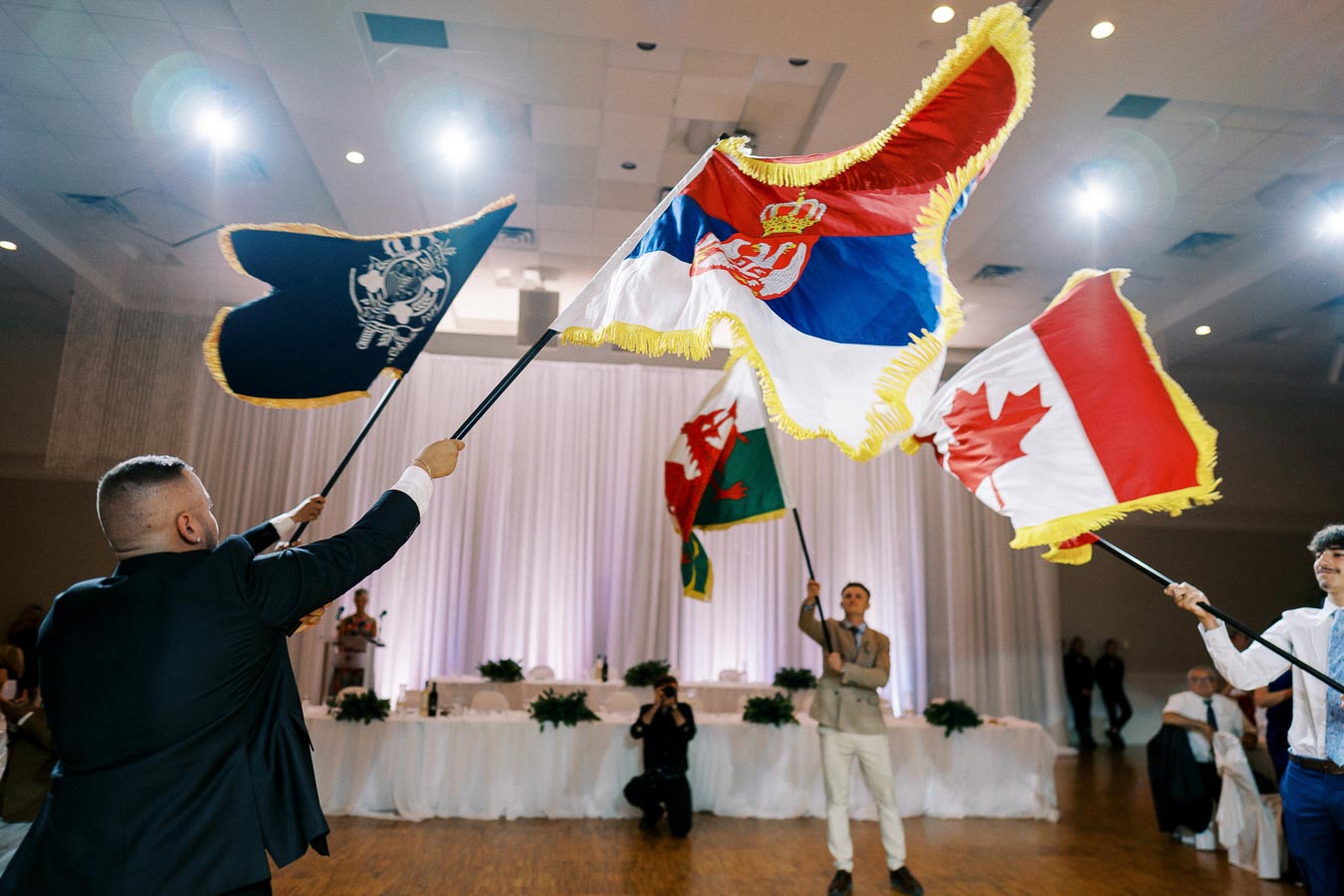 Three men waving Serbian, Canadian, and a black ceremonial flag at an indoor event with a decorated backdrop.