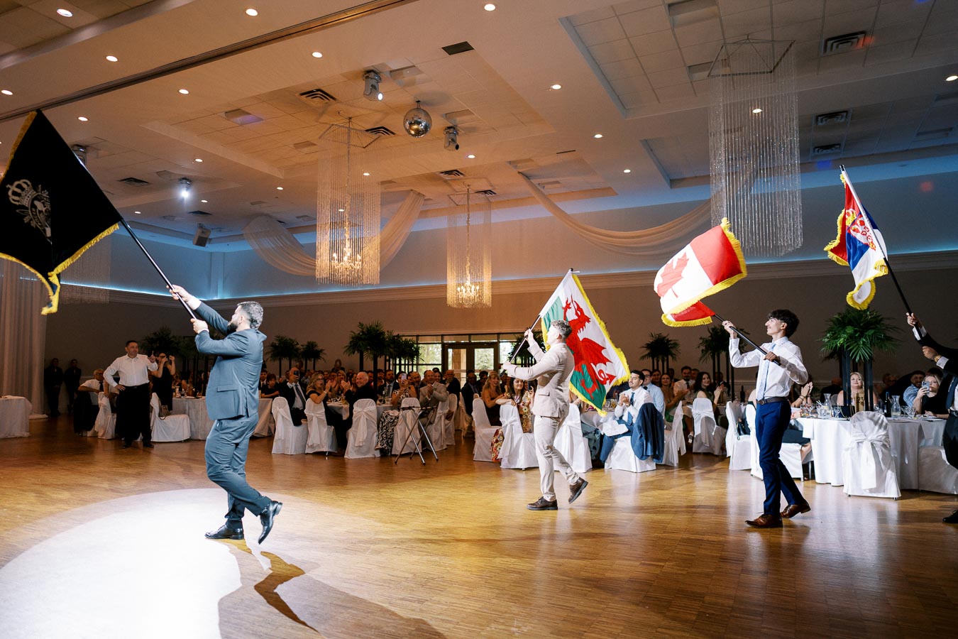A group of people in formal attire marching through a banquet hall, each carrying a different flag, with guests seated at round tables and decorative drapes and chandeliers above.