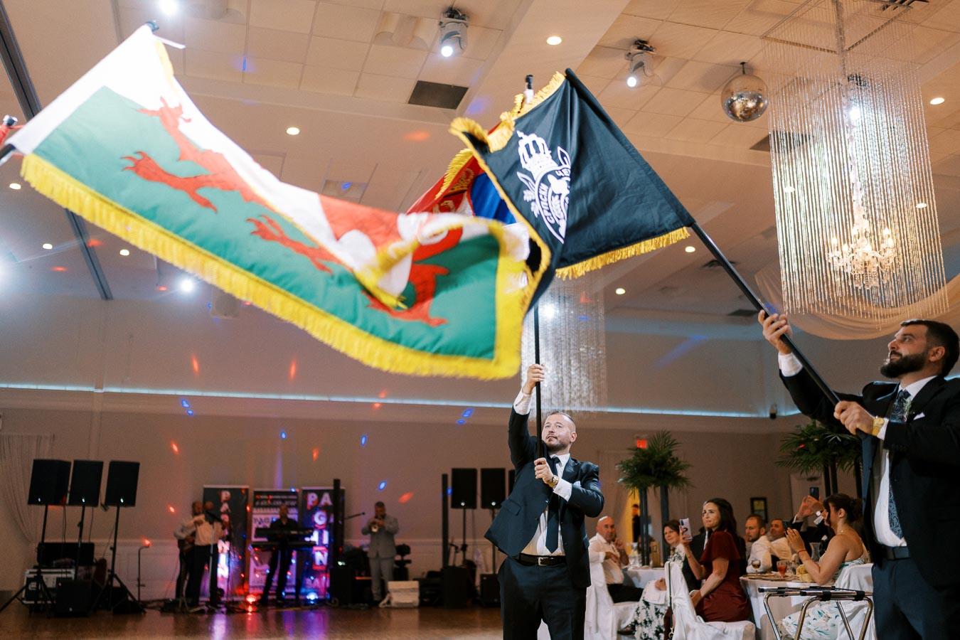 A lively wedding reception scene with elegantly dressed guests watching two men in formal suits waving elaborate flags indoors. The venue is adorned with chandeliers and colorful lights, creating a festive atmosphere.