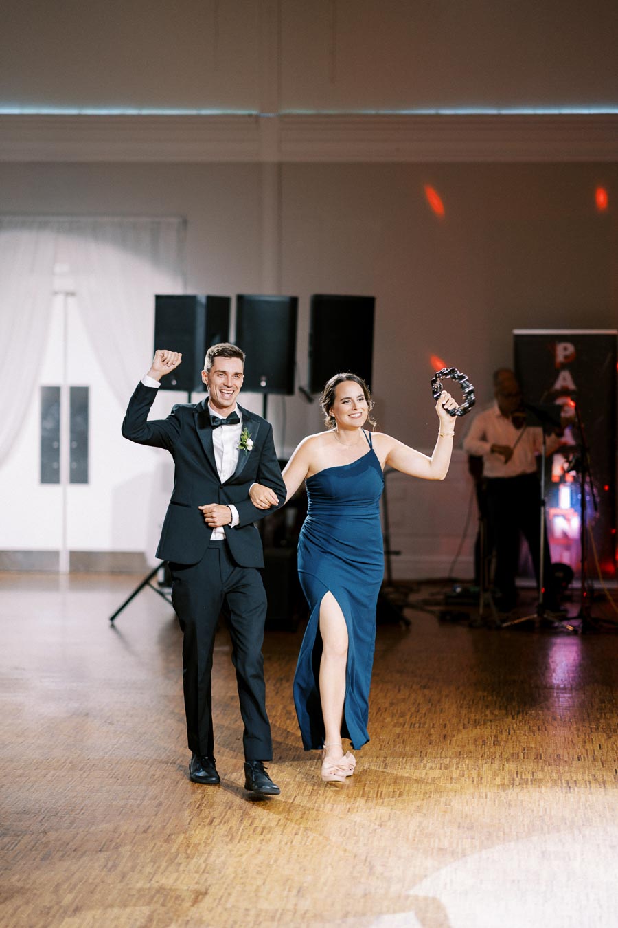 A man in a black tuxedo and a woman in a blue evening gown energetically dance and smile at a formal event, holding a tambourine in a warmly lit indoor venue with a wooden floor and speaker setup in the background.