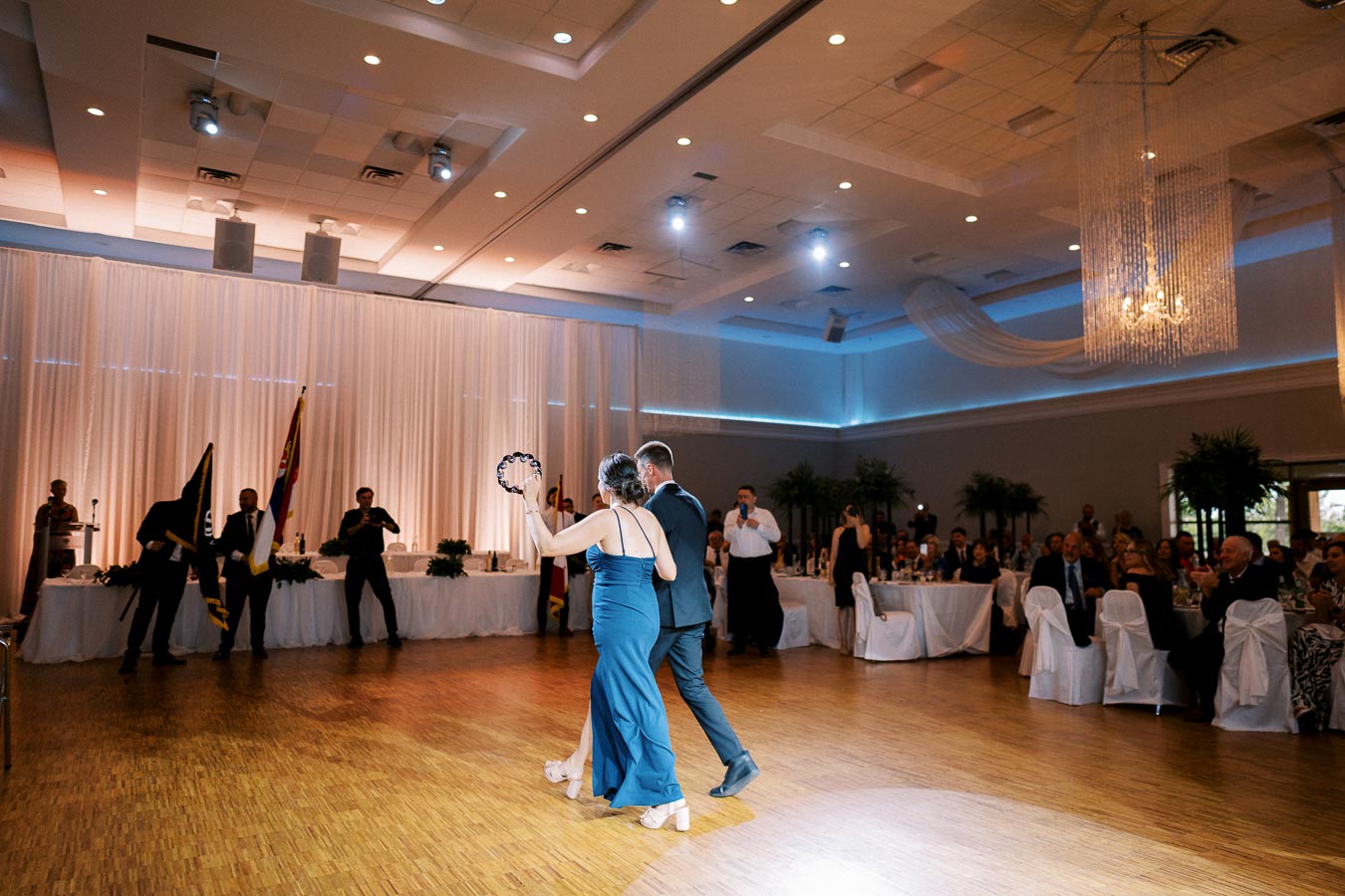 Elegant ballroom with a couple dancing on a polished wooden floor during a formal event, guests seated at round tables adorned with white tablecloths and chairs, and soft ambient lighting enhancing the elegant atmosphere.