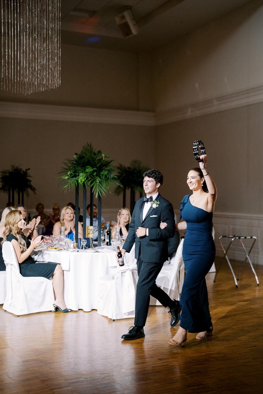 Wedding guests applauding as a man in a tuxedo and a woman in a blue evening gown, holding a tambourine, enter a reception hall with elegantly set tables and lush greenery decor.