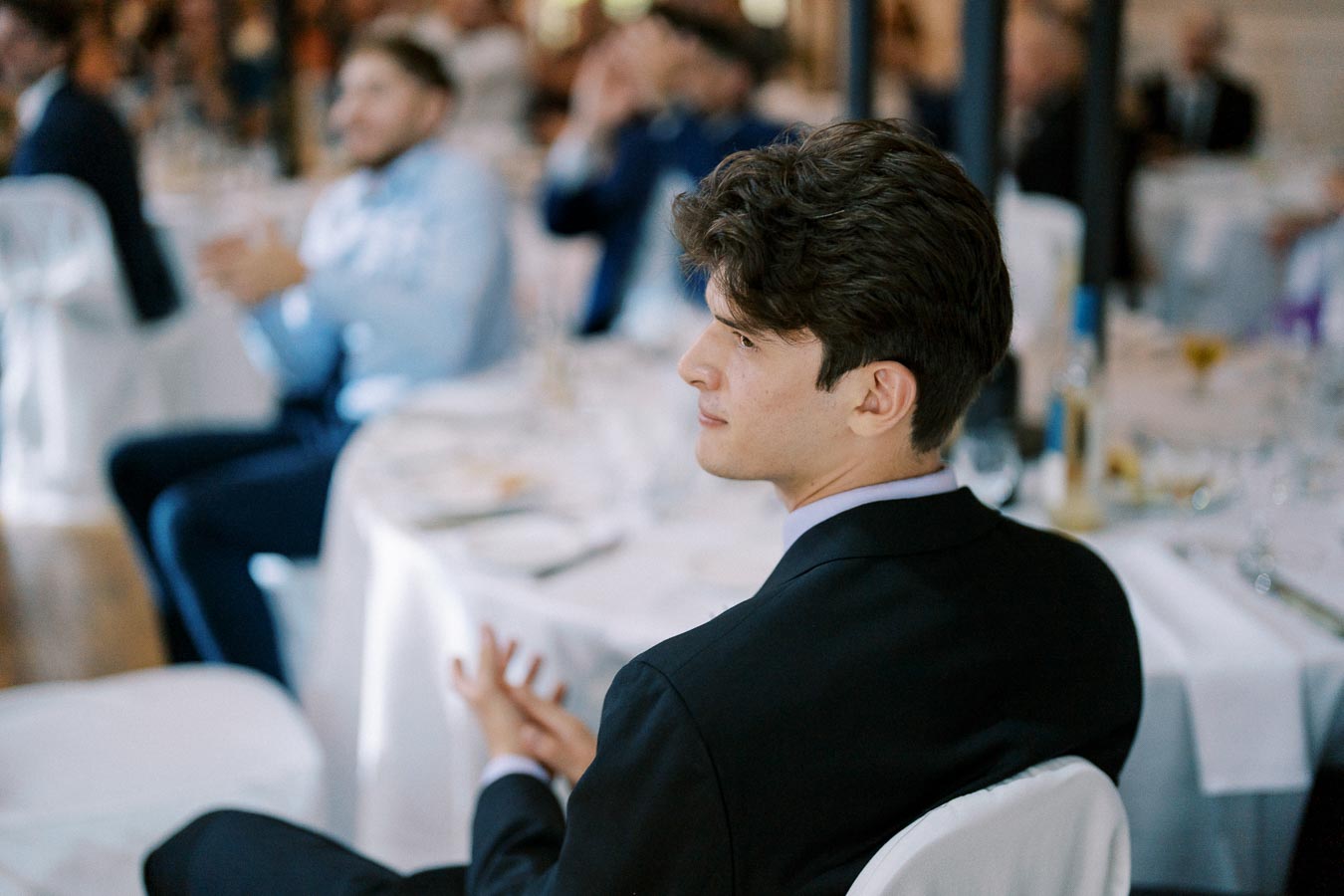 Young man in a formal suit seated at a banquet table, attending an event with elegantly dressed guests in the background.