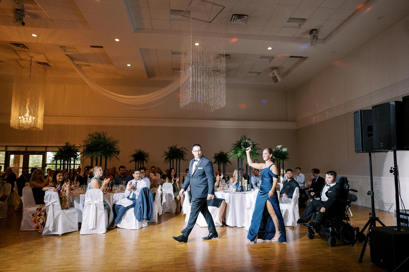 Wedding reception hall with elegantly dressed guests sitting at round tables. A woman in a navy dress and a man in a suit walk in with smiles, while guests look on and applaud. The room features chandeliers, high ceilings, and decorative greenery.