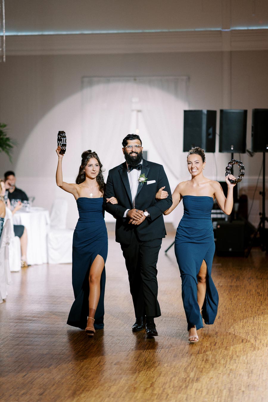 Wedding entrance with two bridesmaids in blue dresses and groomsman in a black suit walking arm-in-arm, holding tambourines, on a wooden dance floor with guests seated at tables in the background.