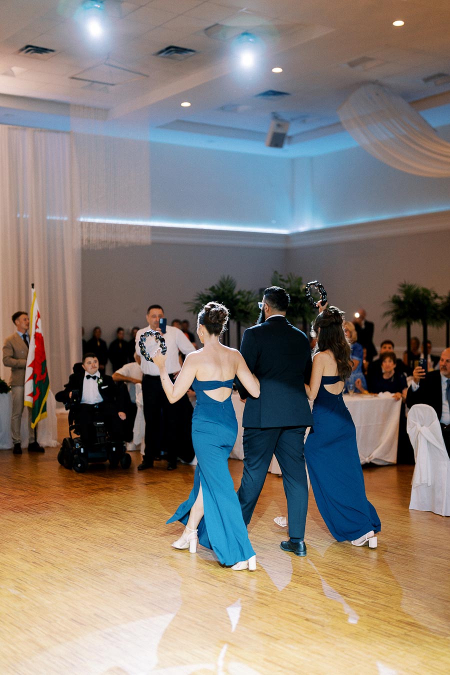 A group of three people in elegant evening attire, holding tambourines, enter a brightly lit banquet hall with guests seated at tables.