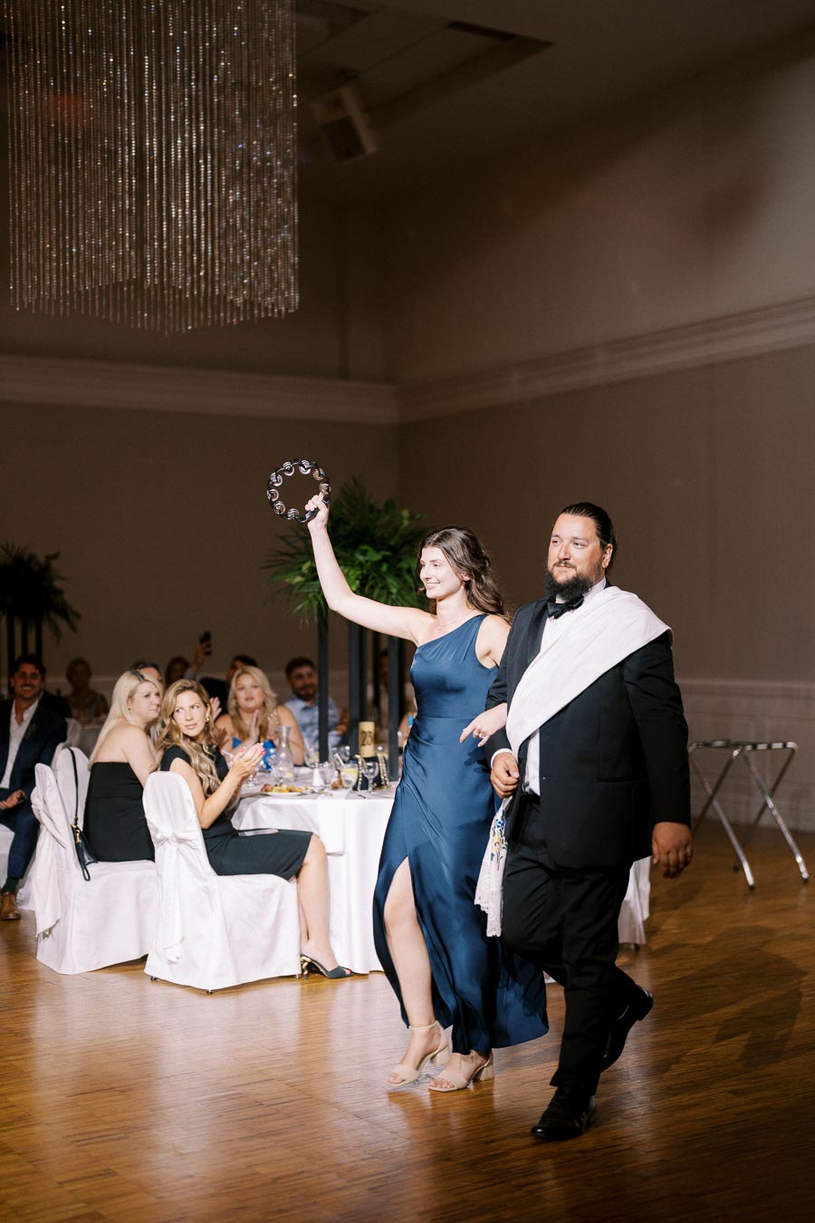 Elegant wedding reception entrance with a woman in a navy blue gown holding a tambourine, escorted by a man in a suit, while guests seated at tables watch and celebrate.