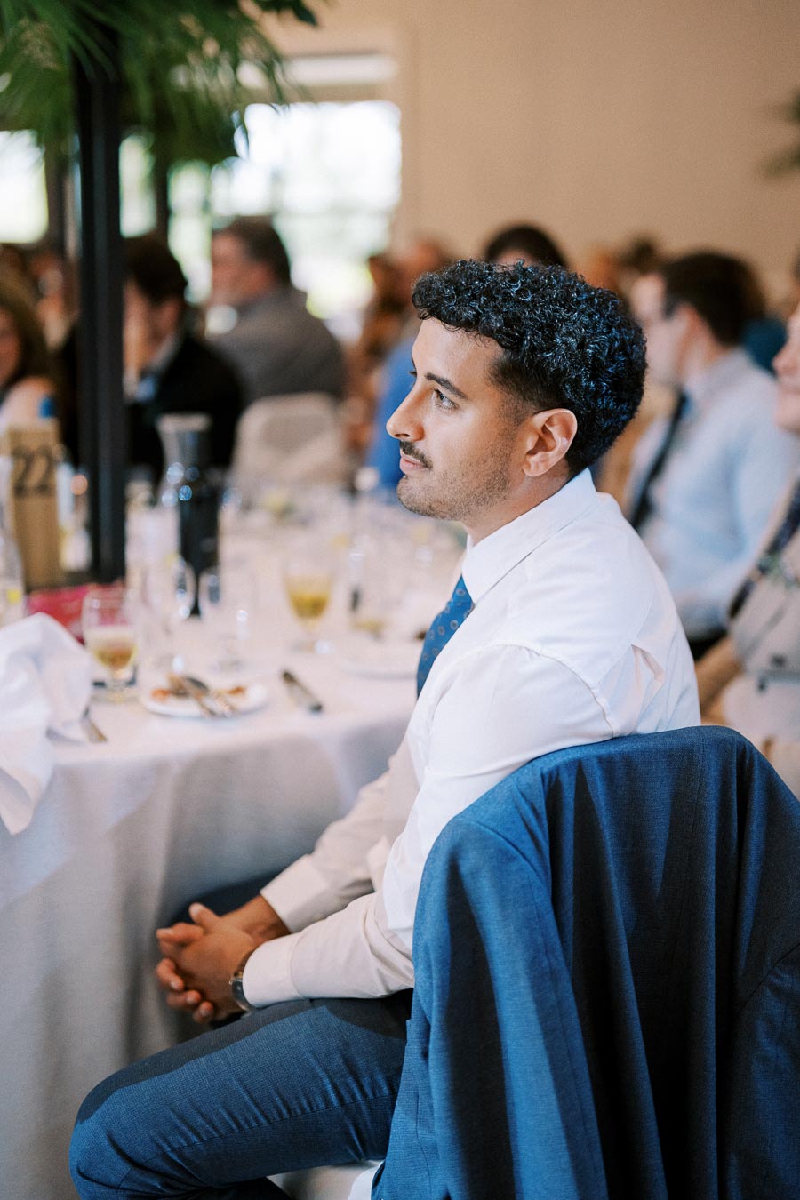 A man in a white shirt and blue suit jacket sitting attentively at a formal event, with a blurred crowd in the background.