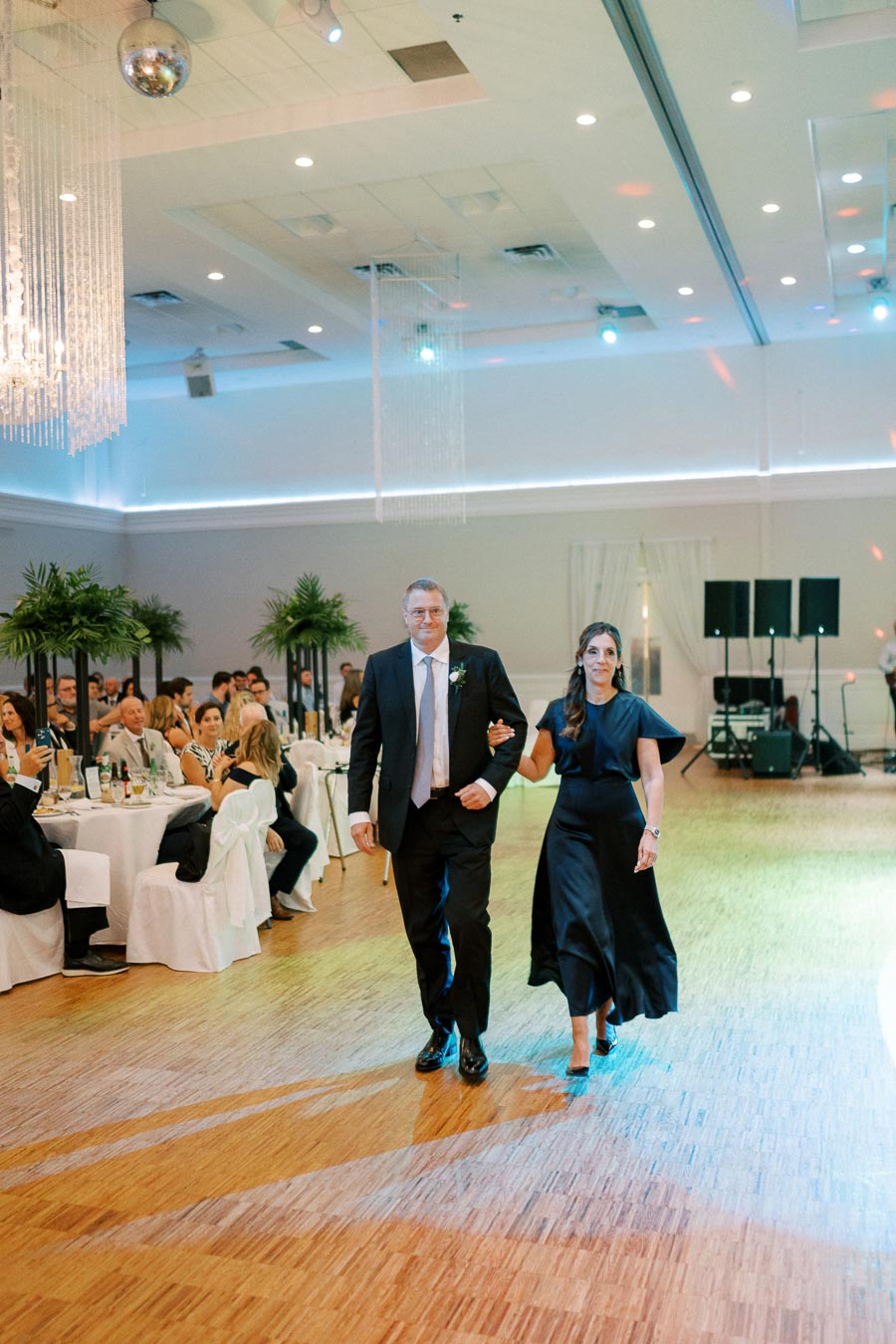 A couple elegantly walking arm in arm on a polished wooden dance floor during a formal event, with well-dressed guests seated at round tables in the background and ambient lighting enhancing the upscale atmosphere.