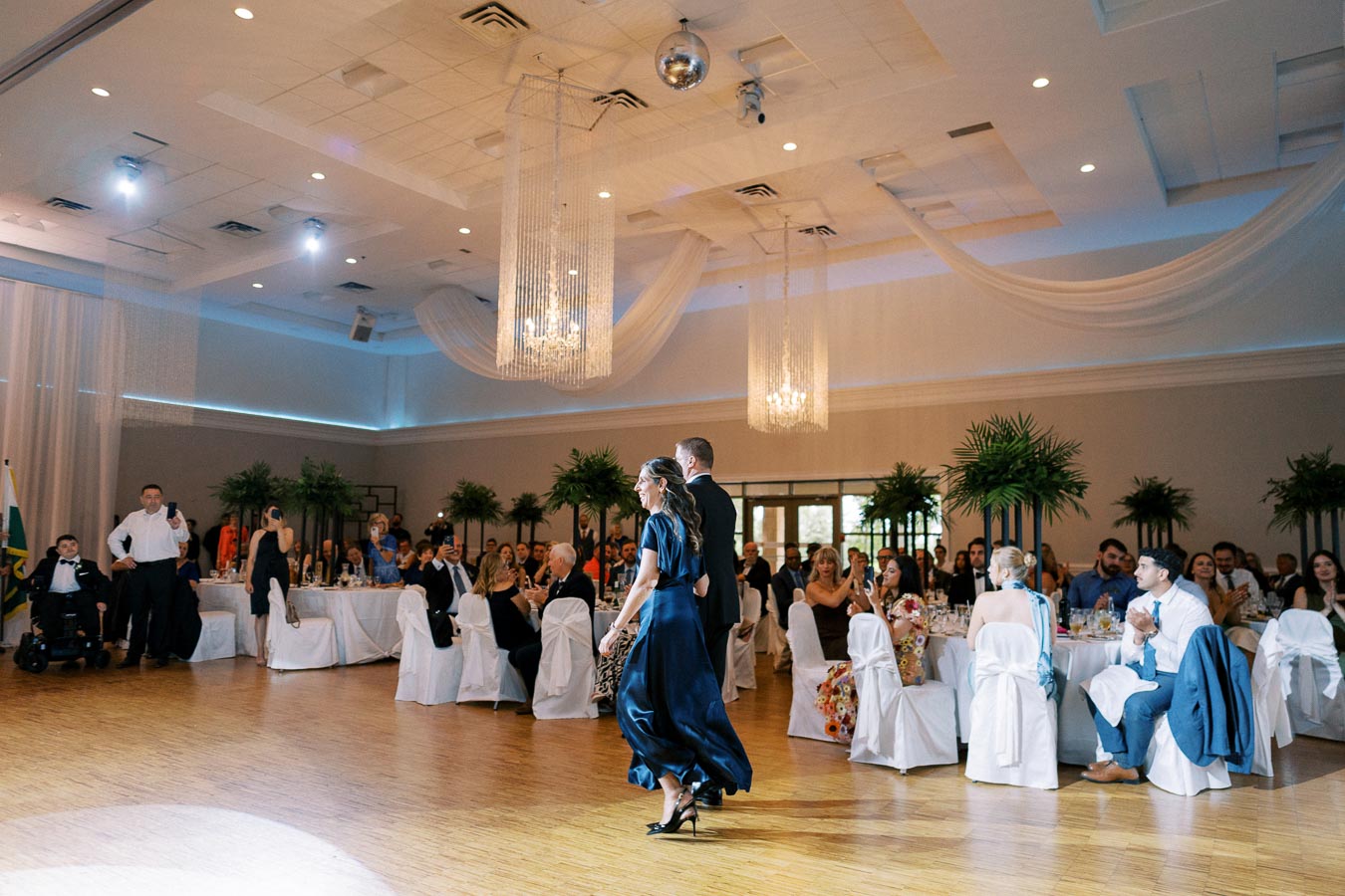 A couple in formal attire walks into a spacious, elegantly decorated banquet hall, filled with seated guests applauding. The room features draped ceilings, ambient lighting, potted plants, and crystal chandeliers, creating a luxurious event atmosphere.