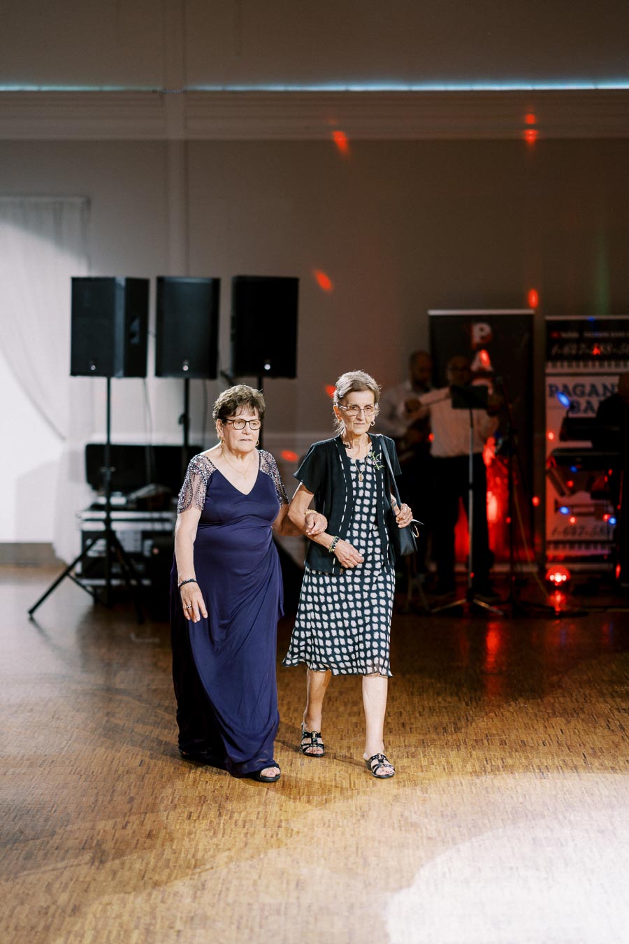 Two women dressed elegantly, one in a blue gown and the other in a black and white dress, walk together arm-in-arm at a formal event, with a band playing in the background.
