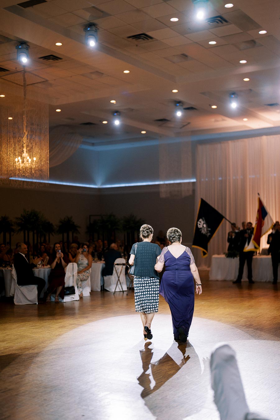 Two elegantly dressed women walk arm in arm on a polished dance floor, surrounded by seated guests in a dimly lit banquet hall during a festive event.