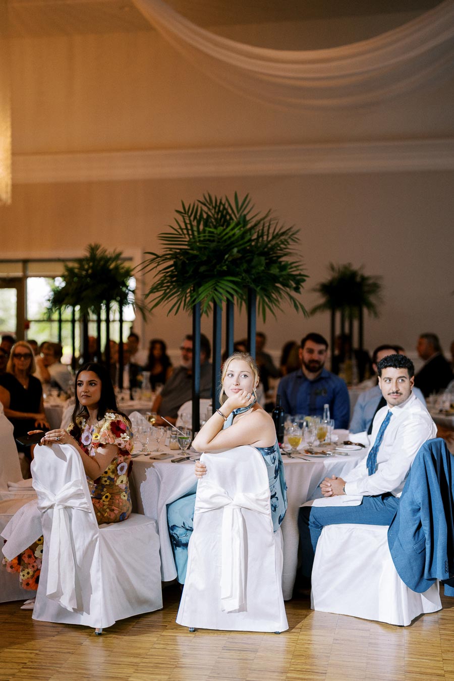 Guests seated at a wedding reception, elegantly dressed and surrounded by white table settings and lush green centerpieces.