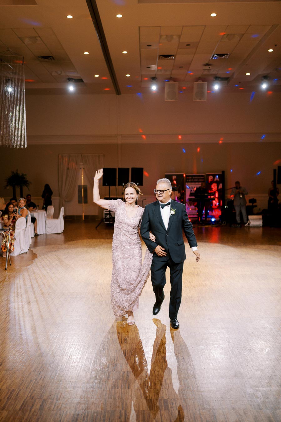 A couple elegantly walking on a polished wooden dance floor at a formal event, with the woman in a sparkling dress waving to the audience and the man in a black tuxedo. Background includes seated guests and a live band under bright event lighting.