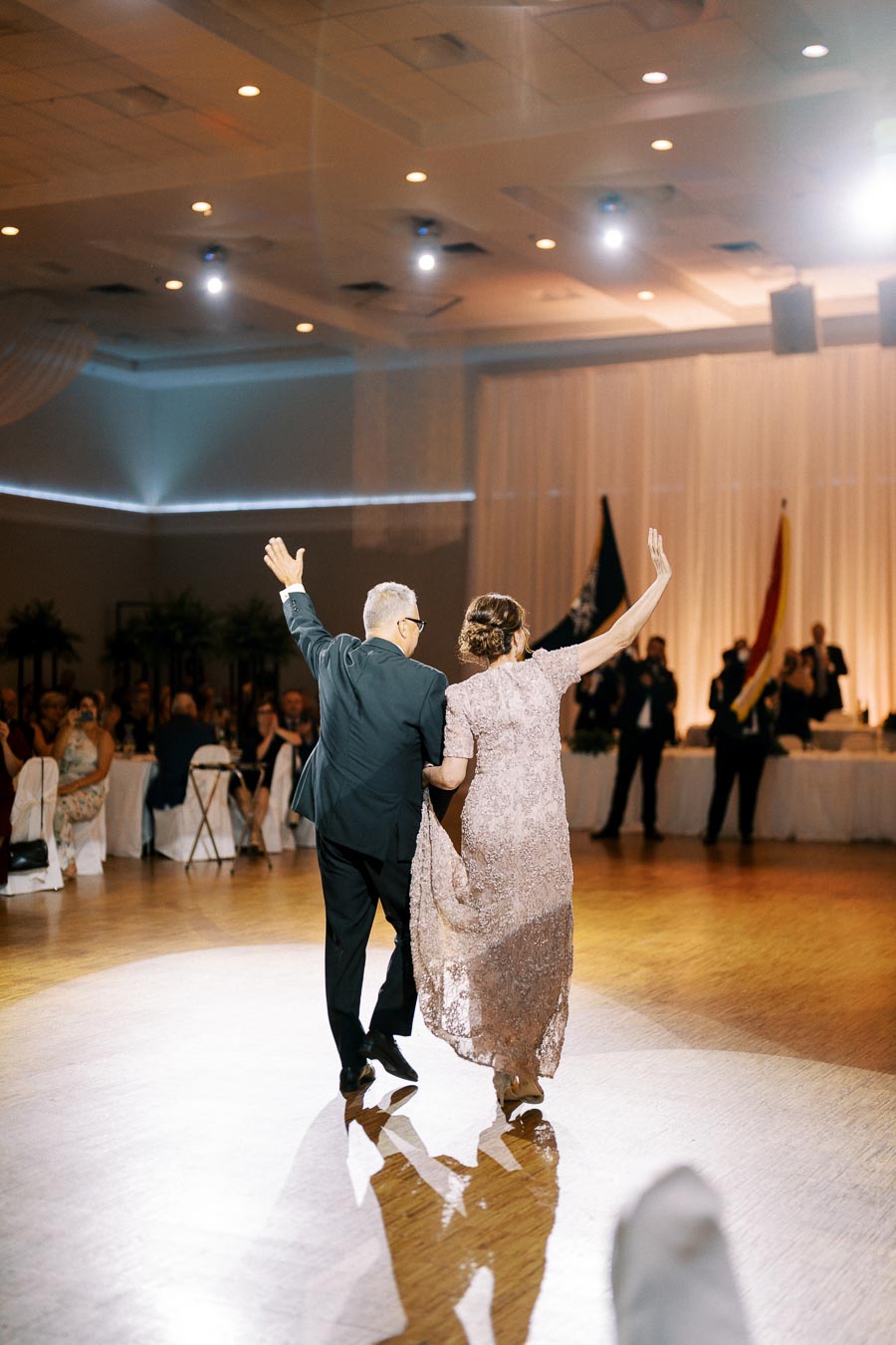 Elderly couple dancing joyfully at a wedding reception in a beautifully lit ballroom, surrounded by seated guests applauding.