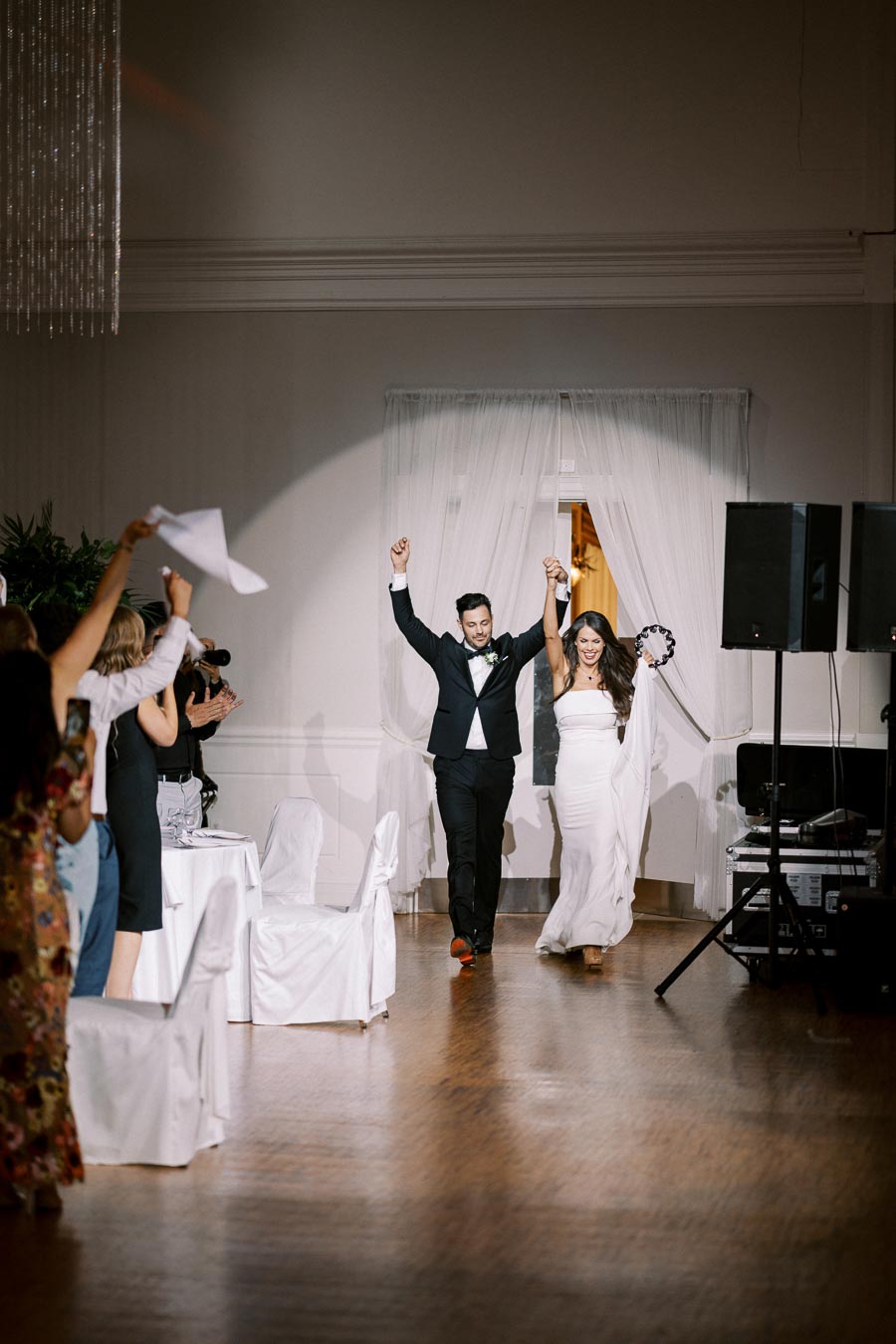 A newlywed couple joyfully enters their wedding reception, holding hands and celebrating as guests cheer around them, in a beautifully decorated venue.