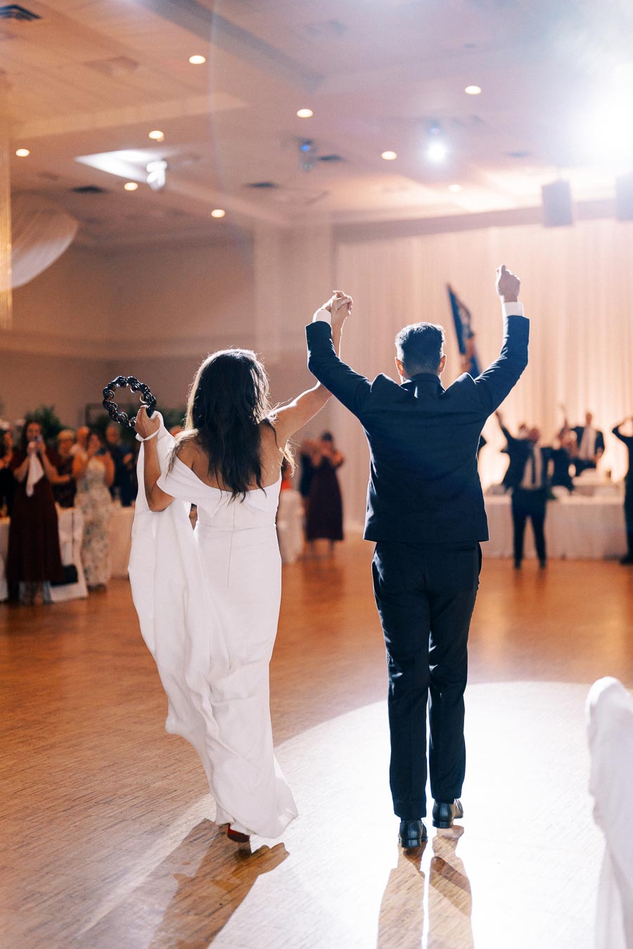 Elegant couple celebrating at wedding reception, bride in white dress holding tambourine, groom in black suit, joyful ambiance with cheering guests and softly lit ballroom