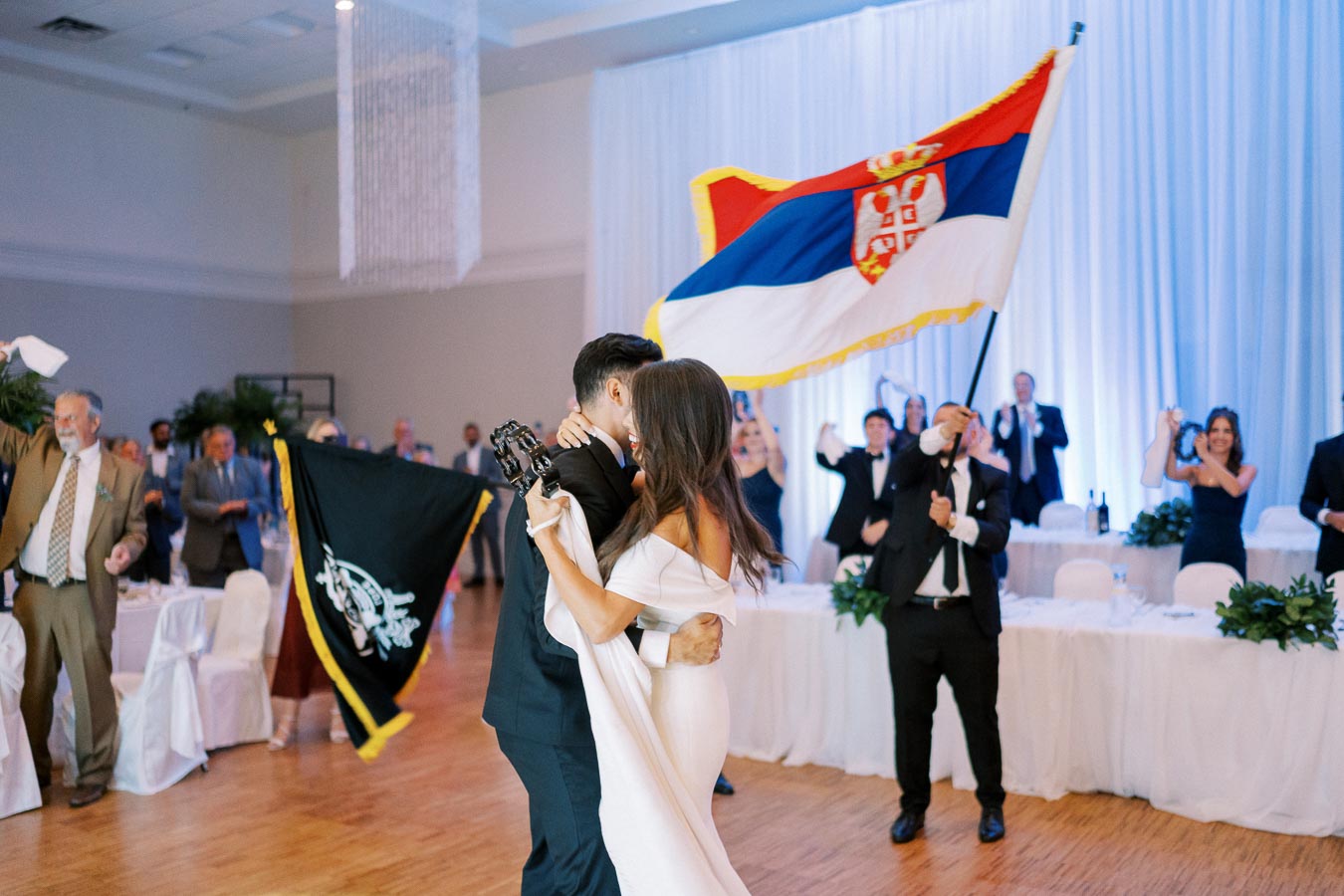 A bride and groom dance joyfully at their wedding reception, surrounded by guests waving flags and cheering. A person holds a large national flag, creating a festive atmosphere in the elegantly decorated venue.