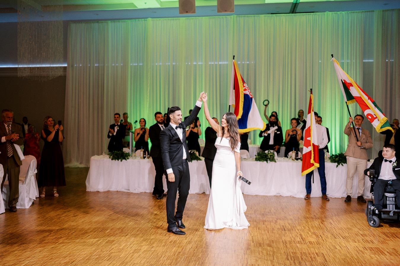 A bride and groom share their first dance at a wedding reception, surrounded by guests holding flags, with a decorated head table and ambient green lighting.