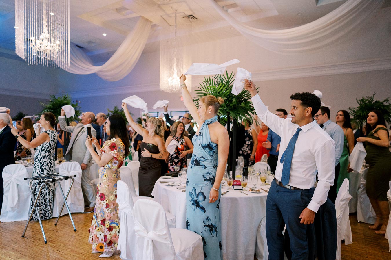 A group of elegantly dressed wedding guests celebrate by waving napkins in a beautifully decorated reception hall with chandeliers and white drapes.