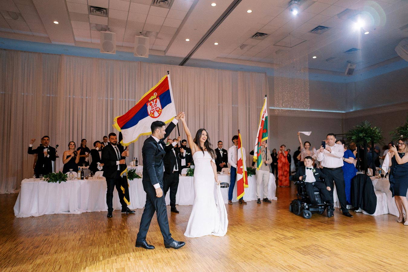 Wedding couple dancing at a reception hall, with the groom holding a Serbian flag. Onlookers, dressed in formal attire, cheer and capture the moment. Various flags are displayed, contributing to the festive atmosphere.