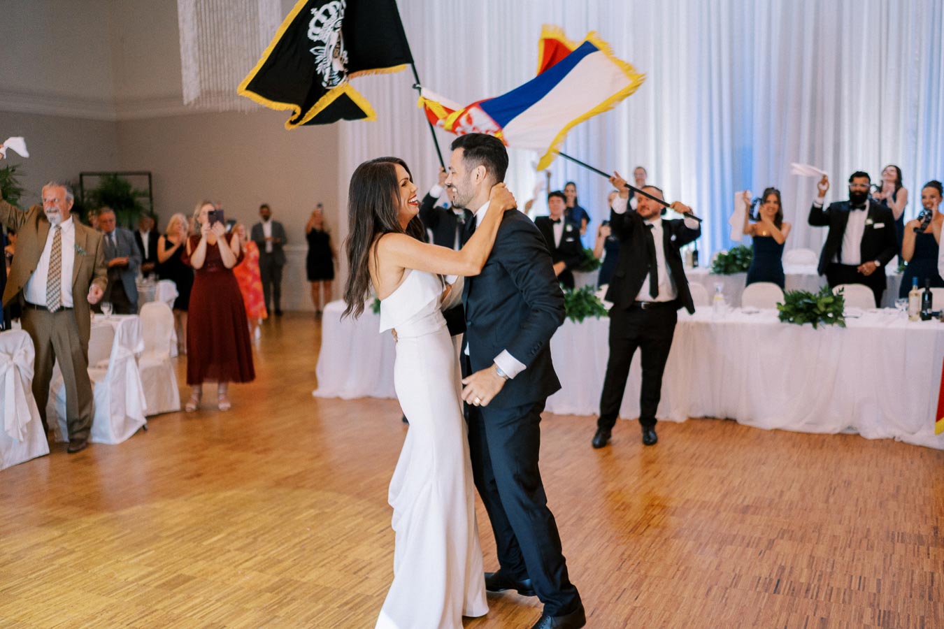 A bride and groom joyfully embrace on the dance floor during their wedding reception, surrounded by cheering guests and vibrant flags.
