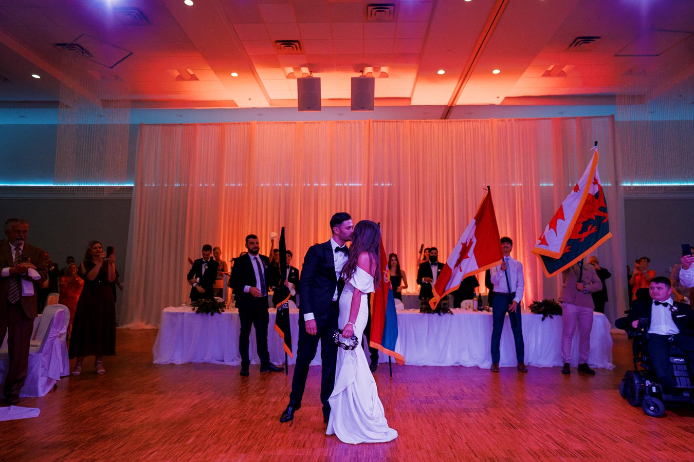 A bride and groom share a kiss on the dance floor during a wedding reception, surrounded by guests holding flags. The room is warmly lit with orange and purple hues, creating a festive and romantic atmosphere.