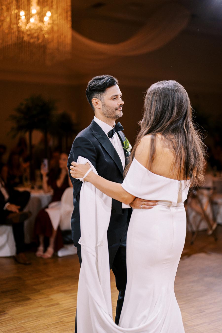 Elegant couple sharing a romantic first dance at a wedding reception, with the groom in a classic tuxedo and the bride in an off-shoulder white gown, under a beautifully lit chandelier.