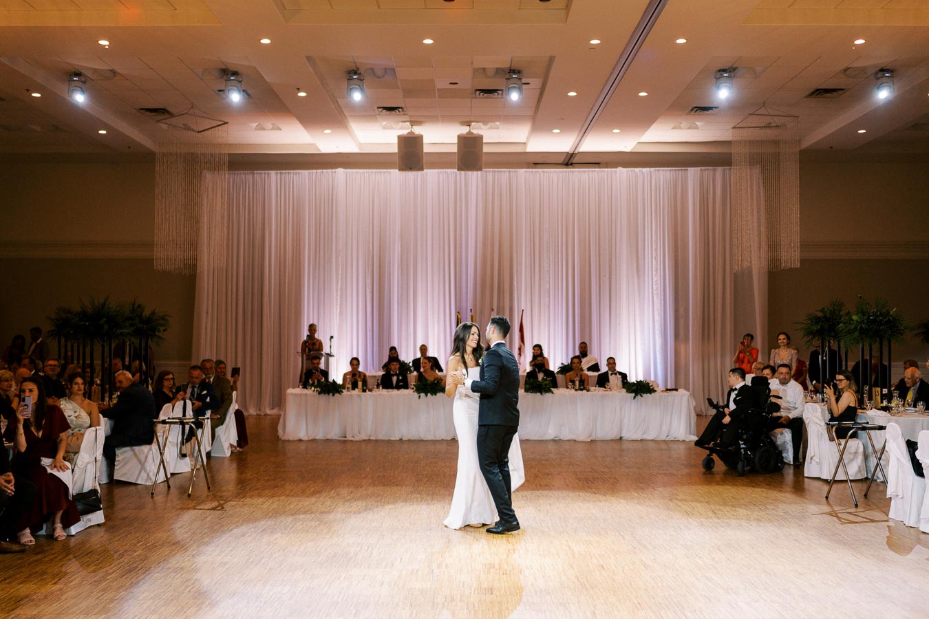Newlywed couple shares their first dance at an elegant wedding reception, surrounded by guests seated at banquet tables, with a backdrop of flowing white drapes and soft lighting.