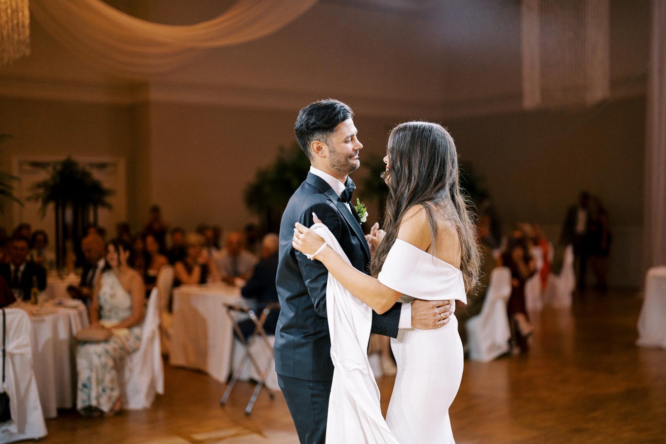 A bride and groom enjoying their first dance at a wedding reception, wearing elegant attire and surrounded by seated guests in a beautifully decorated venue.