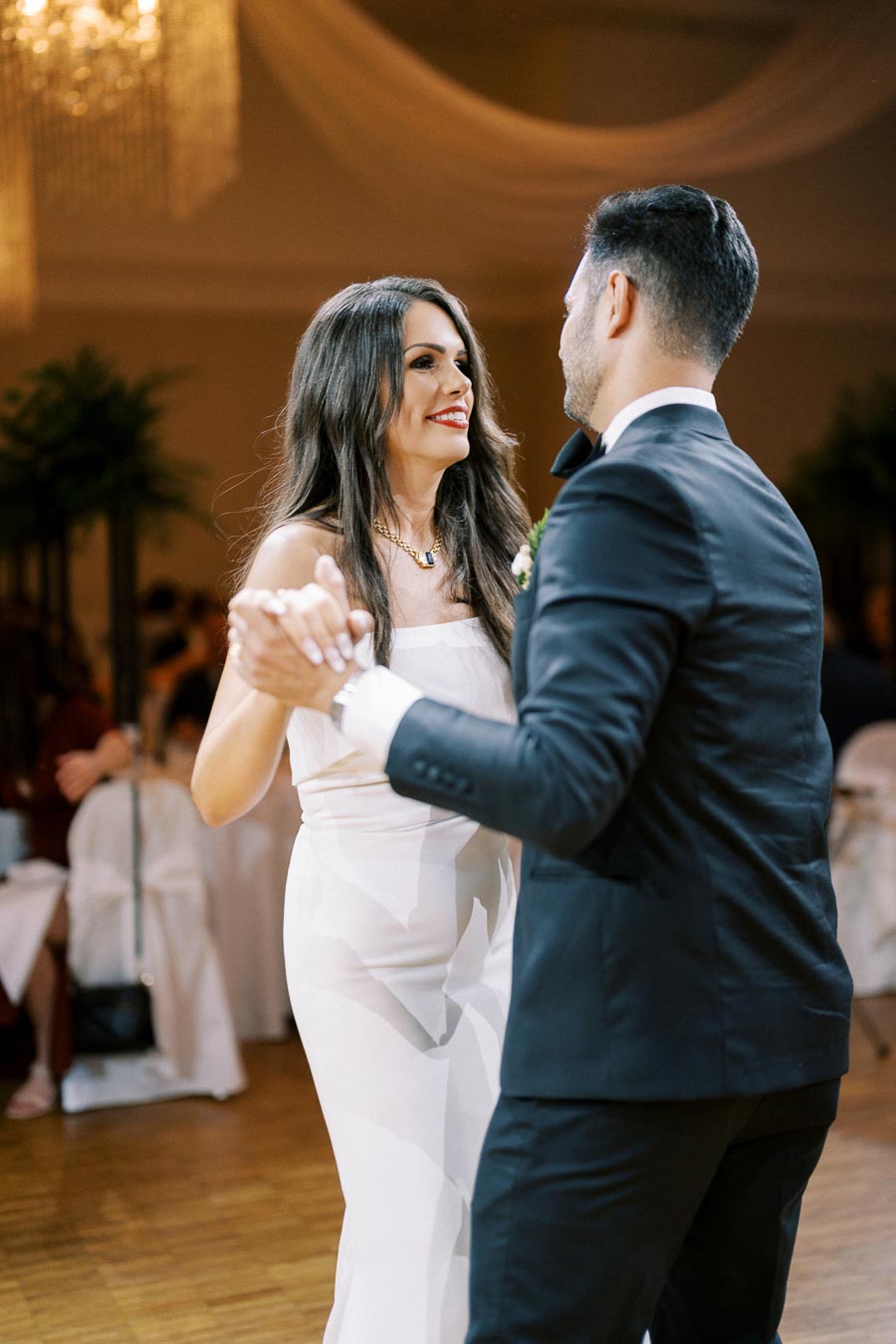 A couple dancing at a wedding reception, with the woman in a white dress and the man in a black suit, elegantly holding each other's hands on a polished wooden floor.