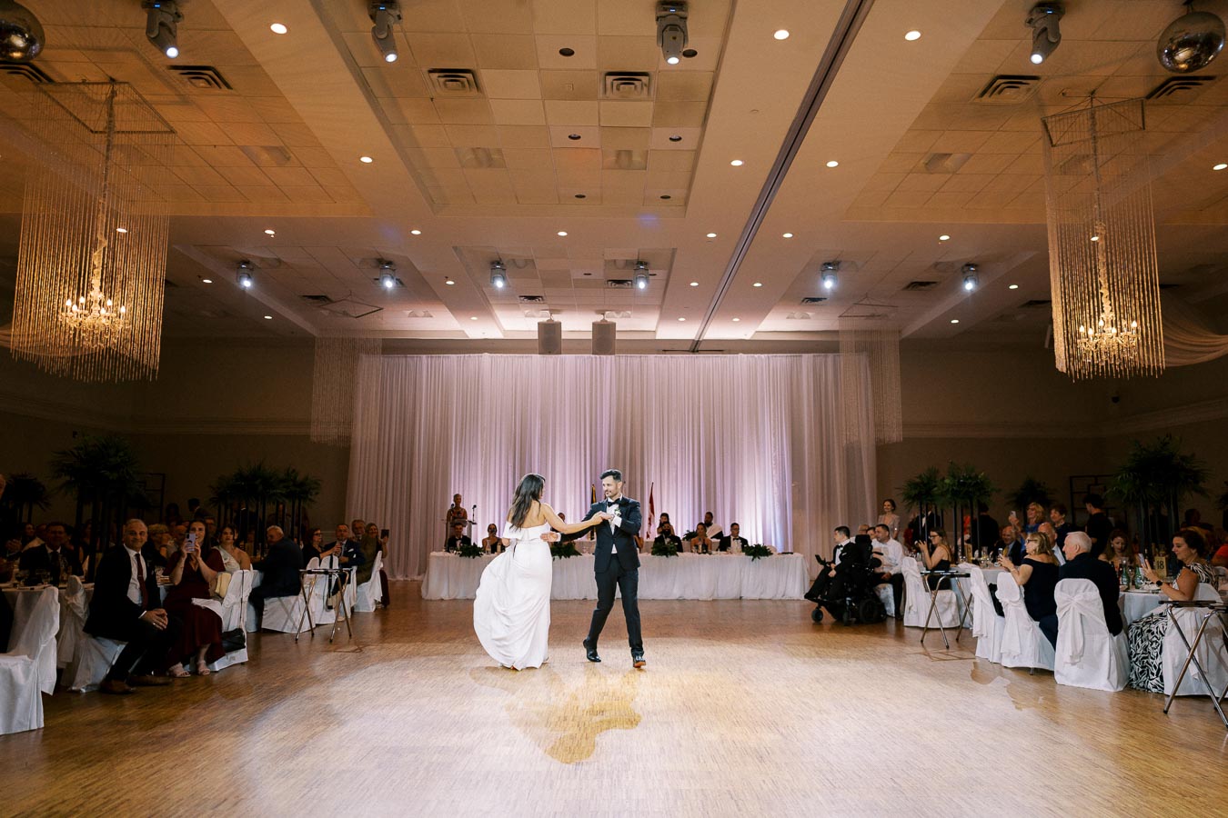 A couple elegantly dancing on a wooden floor in a warmly lit ballroom with guests seated around tables, draped in white linens, and a backdrop of white curtains.