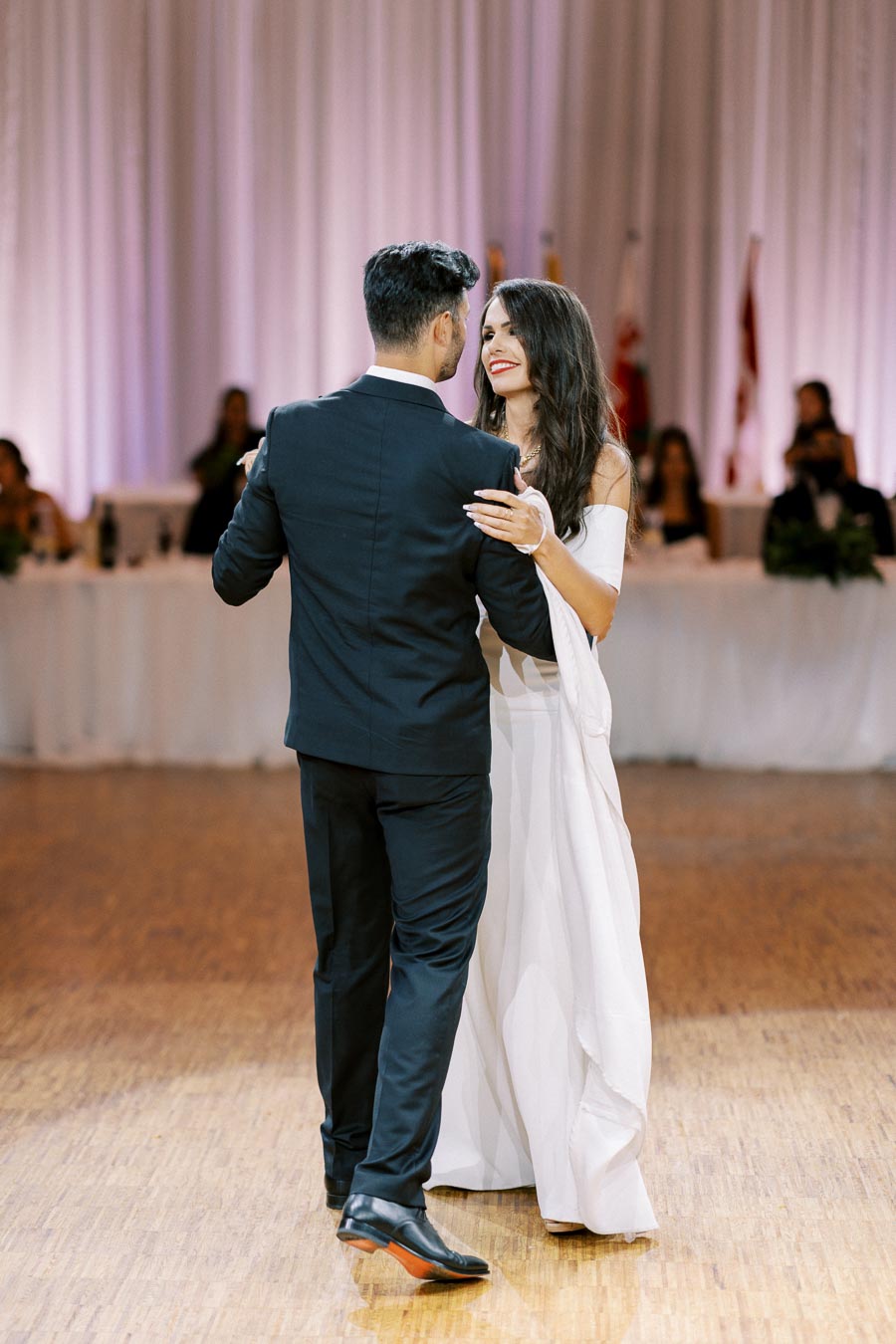 A couple gracefully dancing at a wedding reception, with the woman in an elegant white dress and the man in a black suit, as guests watch in the background.