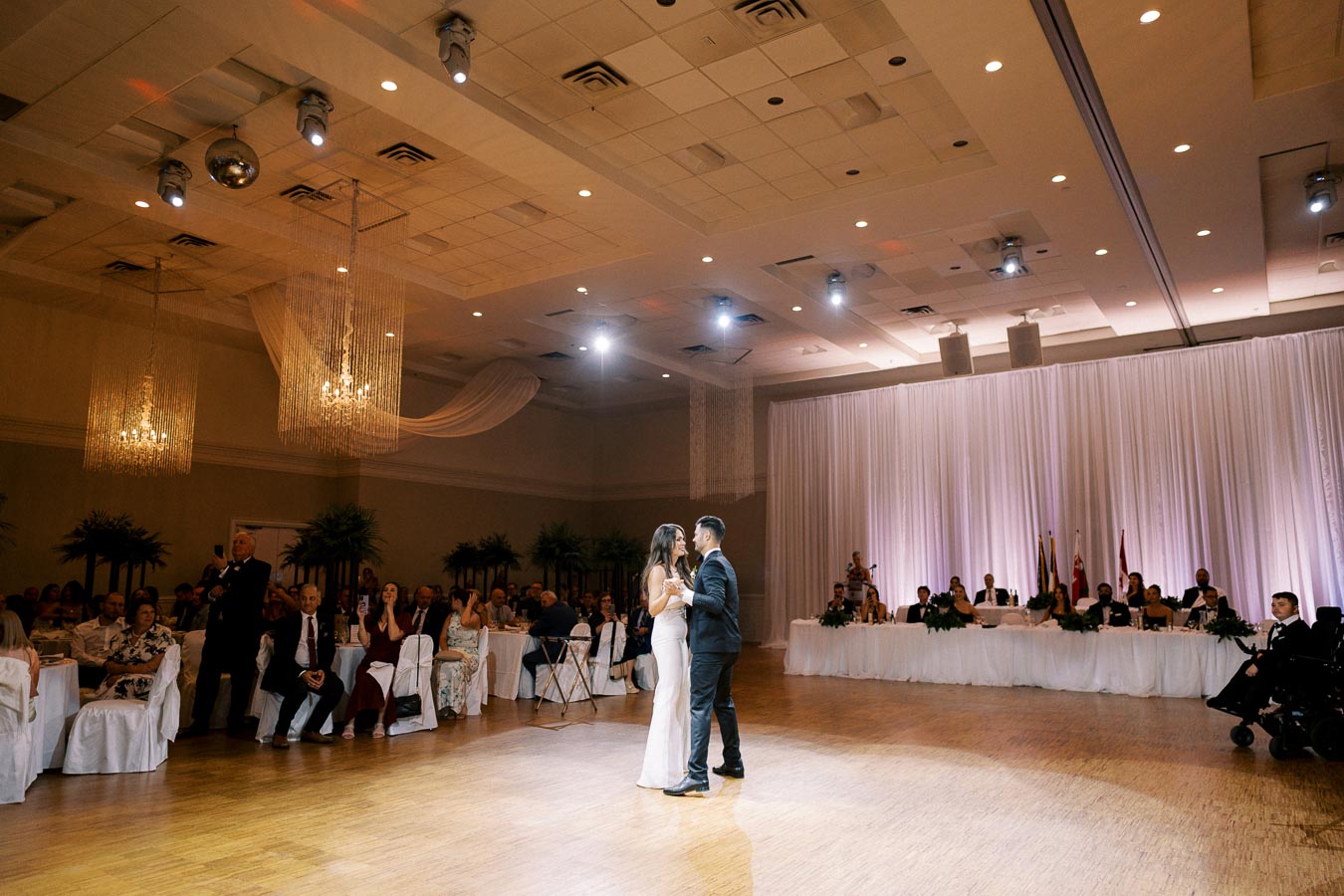 A couple shares their first dance at a wedding reception in a beautifully decorated ballroom. The room features elegant chandeliers, draped white curtains, and warmly lit ambiance. Guests are seated around the dance floor, attentively watching the couple.