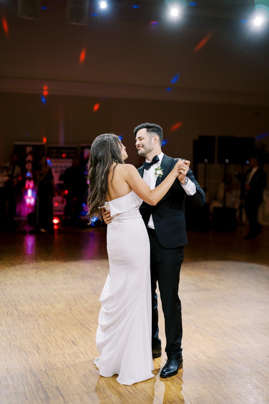 Couple sharing their first dance at a wedding reception, elegant white gown and black tuxedo, romantic celebration atmosphere with soft lighting and colorful reflections on the dance floor.