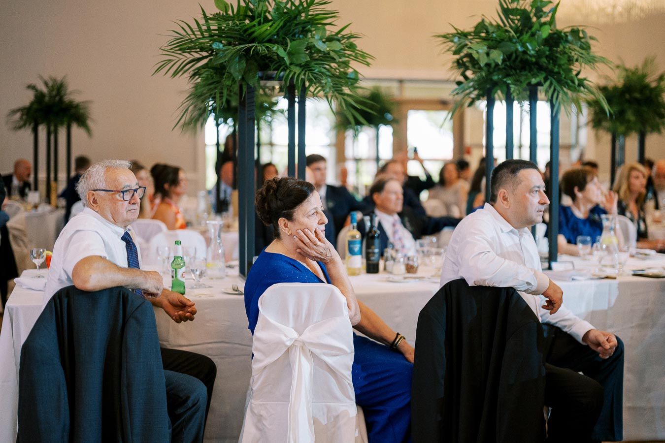 Guests seated at elegantly decorated wedding reception, attentively listening to a speech, with lush green centerpieces on tables.