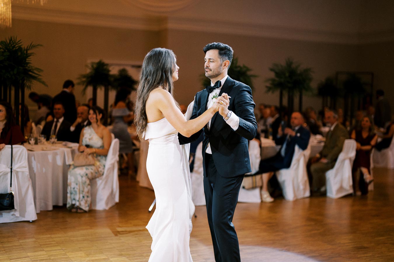 A couple elegantly dances at a wedding reception, surrounded by seated guests in a warmly lit banquet hall.