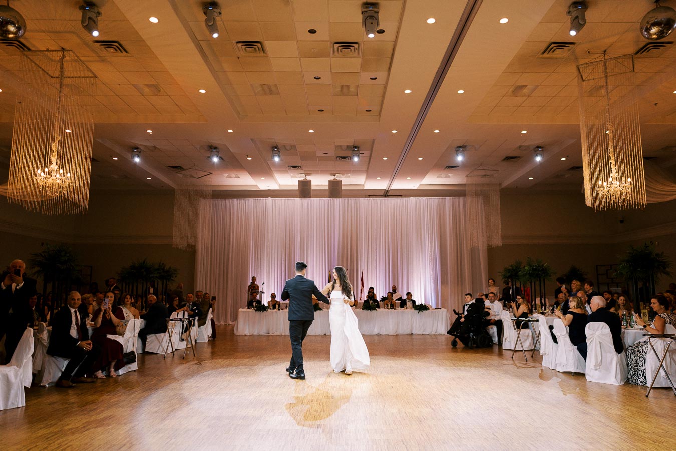 A bride and groom share their first dance at a wedding reception, surrounded by seated guests in an elegantly decorated ballroom under soft ambient lighting.