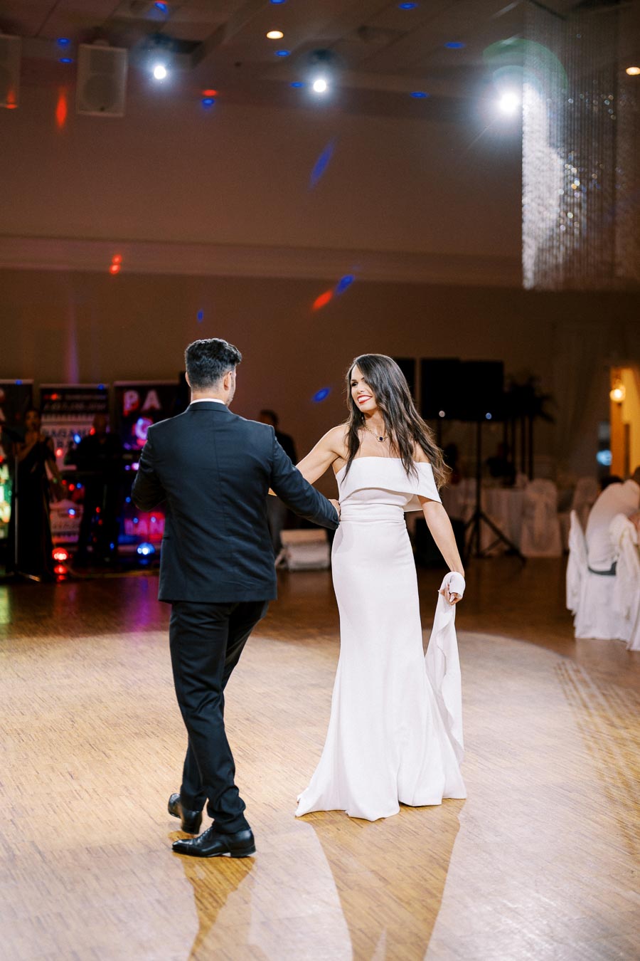 A couple elegantly dancing on a wooden floor in a dimly lit ballroom, with the woman wearing a white dress and the man in a black suit, surrounded by lights and a festive atmosphere.