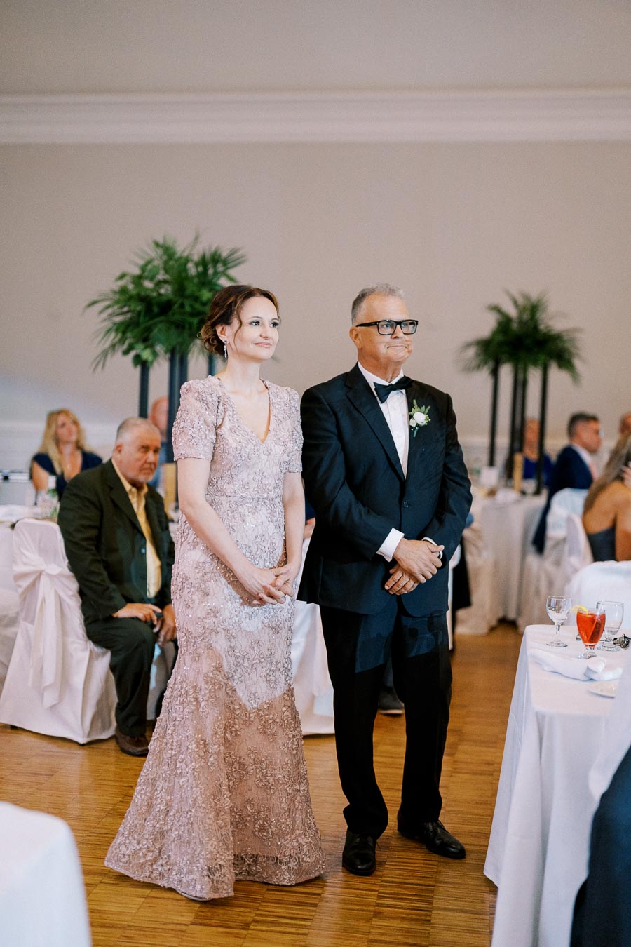 Elegant couple in formal attire standing at a wedding reception, with decorated tables and guests in the background.