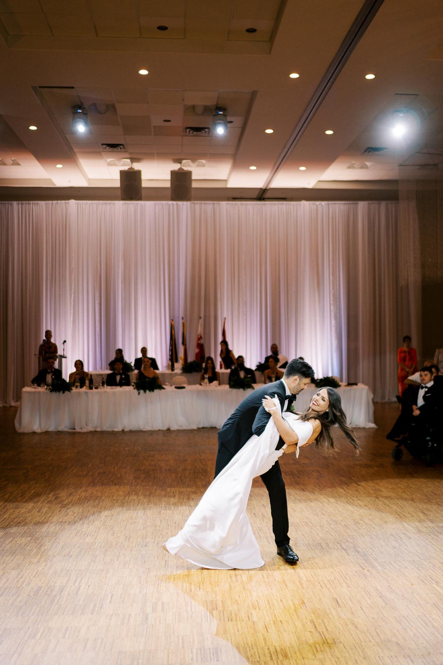 A bride and groom perform their first dance at a wedding reception, surrounded by elegantly draped curtains and an audience seated at a long table. The couple is captured mid-dip on a polished wooden dance floor under soft overhead lighting.