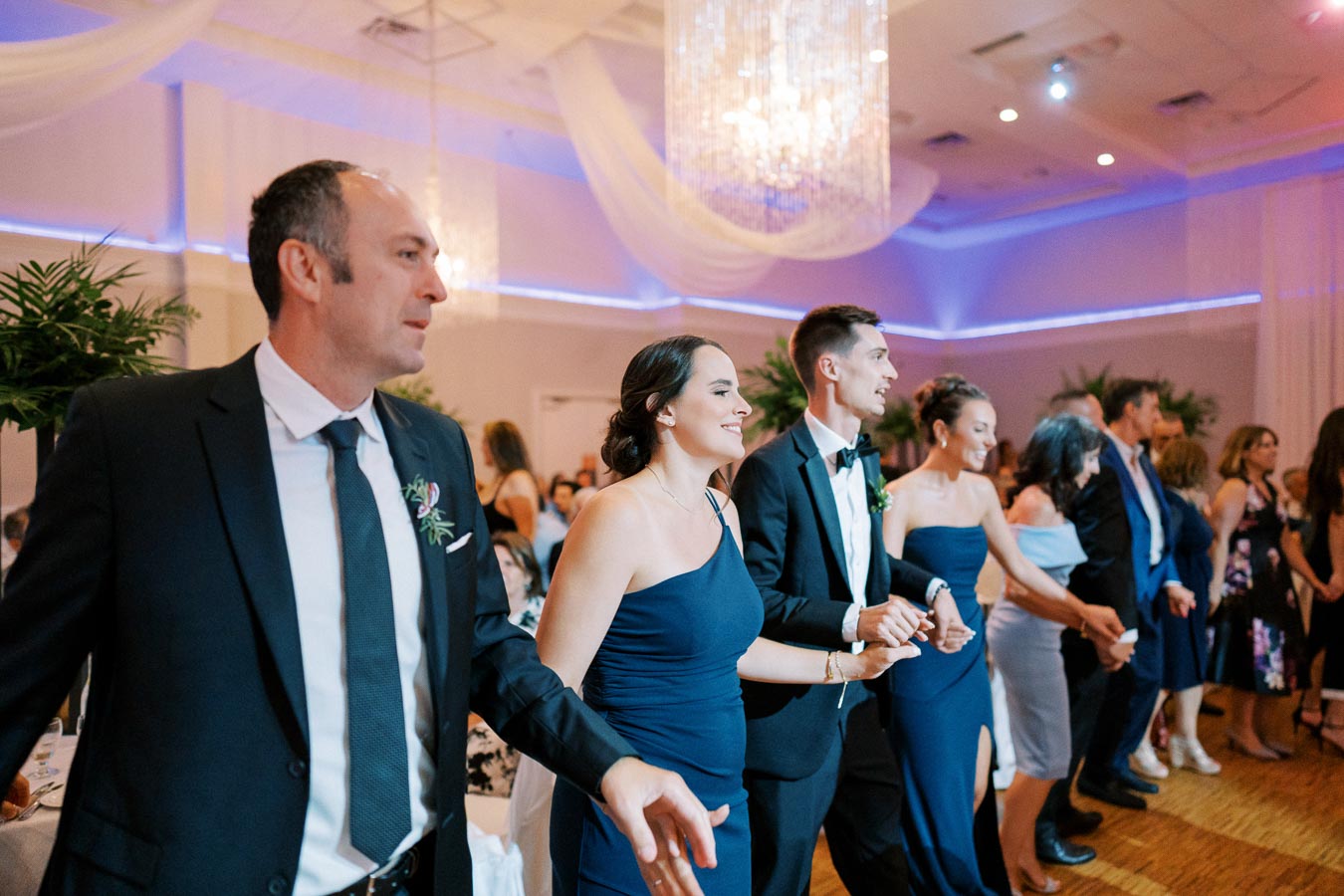 Group of elegantly dressed people holding hands and dancing at a formal wedding reception with chandeliers and festive lighting.