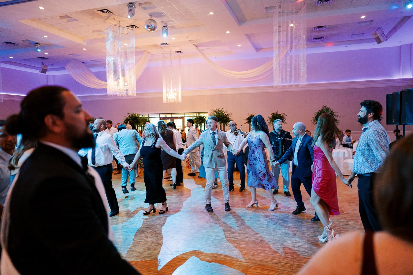 A group of people holding hands and dancing in a circle at a lively indoor wedding reception, with elegant chandeliers and draped fabric hanging from the ceiling, creating a festive atmosphere.