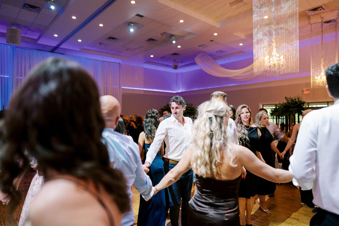 Group of people dancing at a wedding reception in an elegant venue with chandeliers and ambient blue lighting.